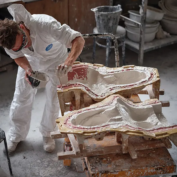 A view in the workshop. The two parts of the mold are placed on a workbench. Our artisan, in a white jumpsuit, is applying a first coat of plaster with a fine brush in one of the molds