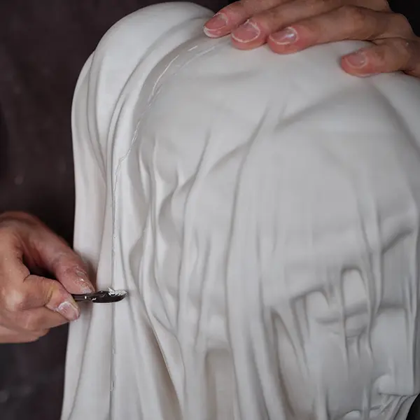 Close-up on the Veiled Virgin. We see the hand of the artisan, holding a scalpel, trimming the cast